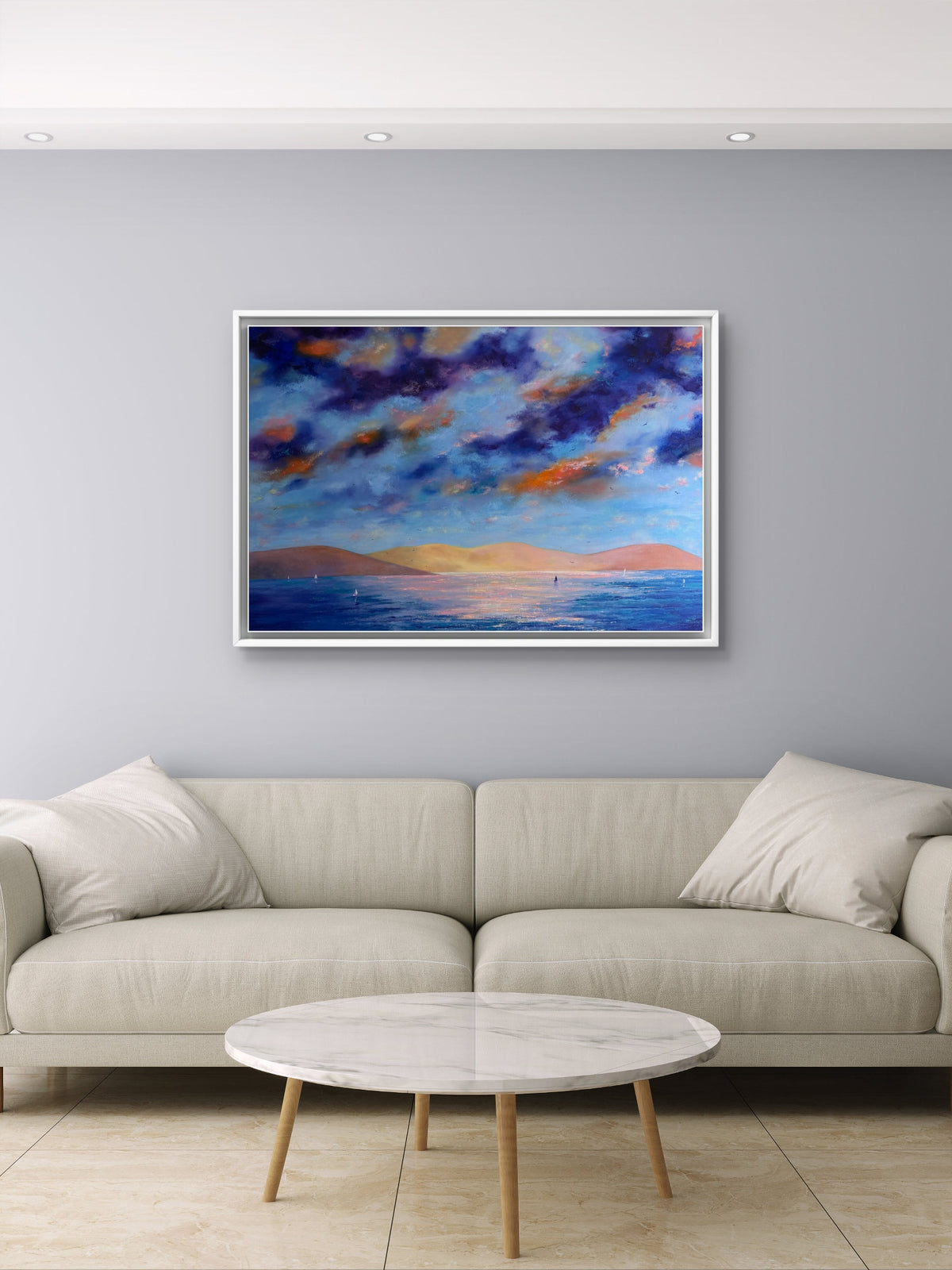 A framed landscape painting of a dramatic blue and orange sunset sky over water, displayed on a light gray wall above a modern beige sofa and round marble coffee table.
