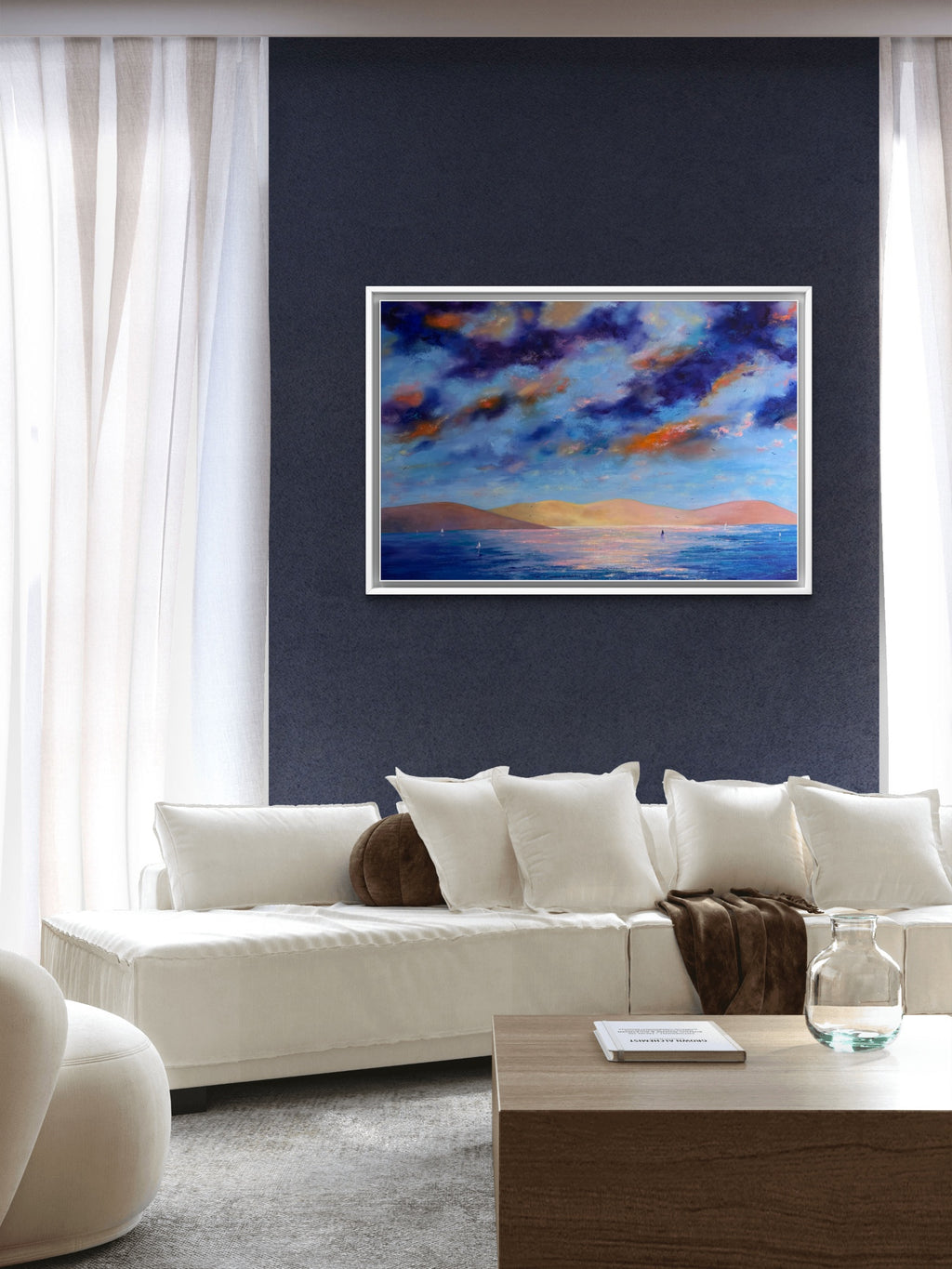 A framed square painting of a colorful sunset over water hangs on a dark navy wall above a modern white sectional sofa in a bright living room.