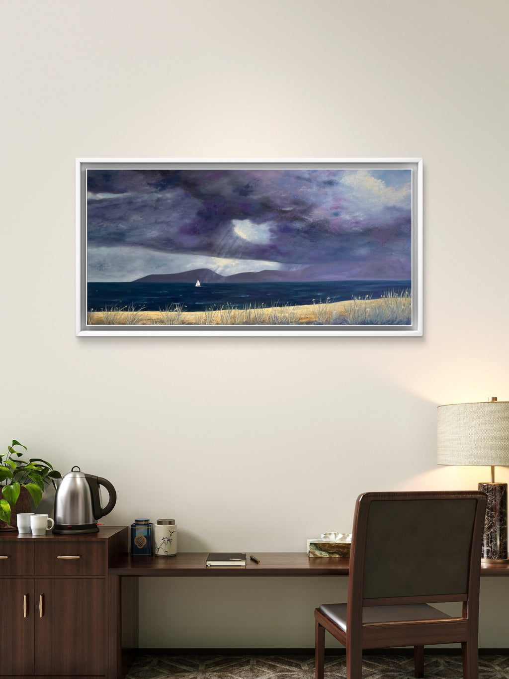 A framed landscape painting of a stormy sea with dark purple and blue clouds, a small sailboat, and a pale sandy shore, displayed on a beige wall above a wooden desk with a chair, kettle, and lamp.
