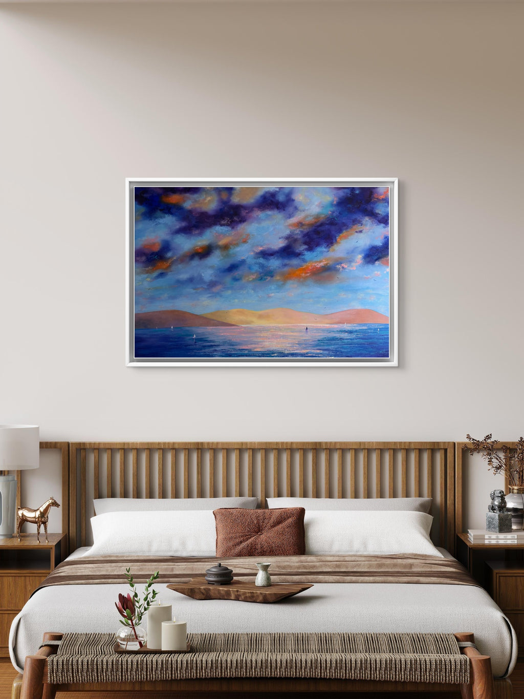 A framed landscape painting of a dramatic blue and orange sunset sky over water, displayed on a beige wall above a wooden bed in a modern bedroom setting.