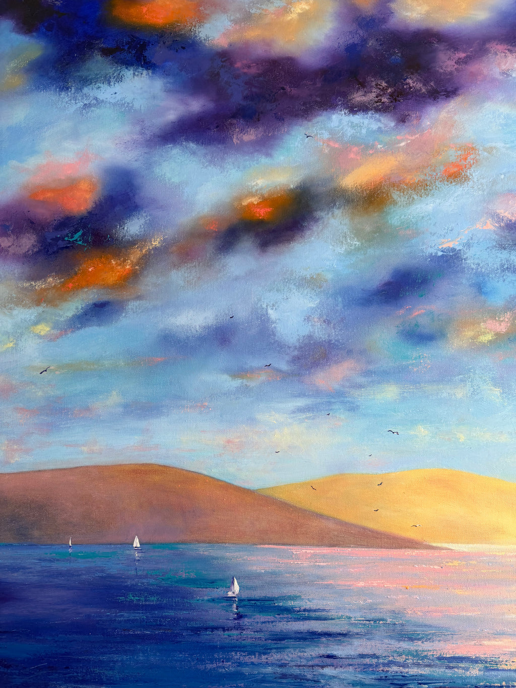 An impressionistic landscape painting of a seascape under a dramatic cloudy sky. The scene features deep blue water with small sailboats, golden-yellow hills on the right, and a textured sky filled with shades of blue, purple, orange, and pink.