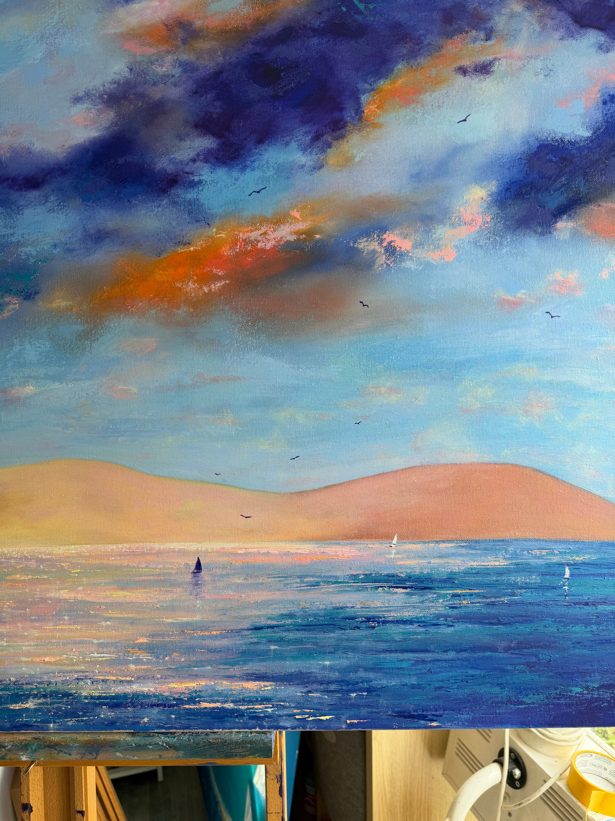 An impressionistic landscape painting of a seascape under a dramatic cloudy sky, with blue water, soft pink hills, and deep blue and orange clouds.