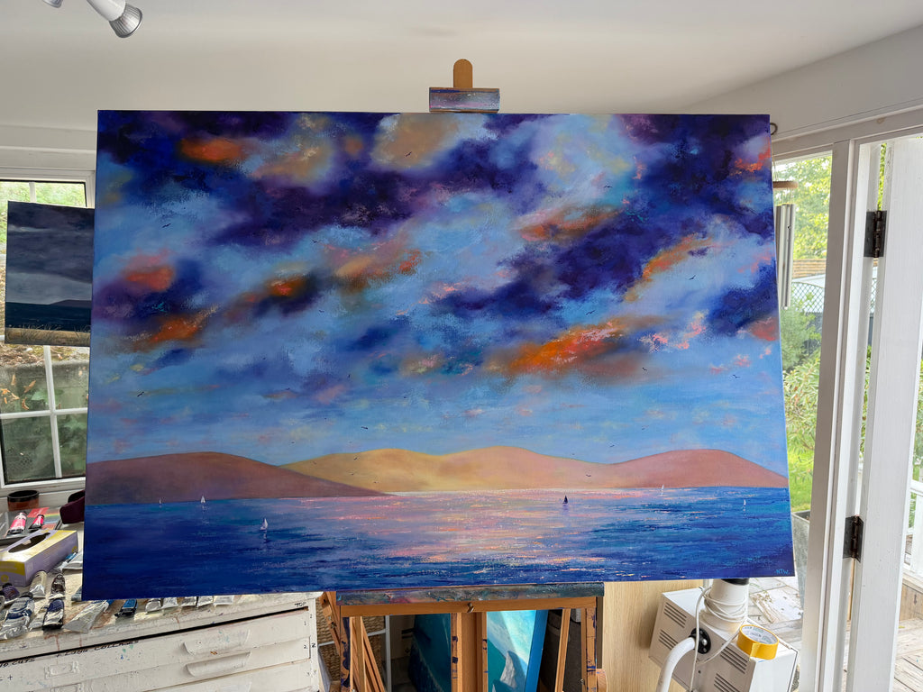 A large canvas painting of a seascape with dramatic blue and purple clouds, orange highlights, and blue water with low tan hills, displayed on an easel in a bright artist studio.