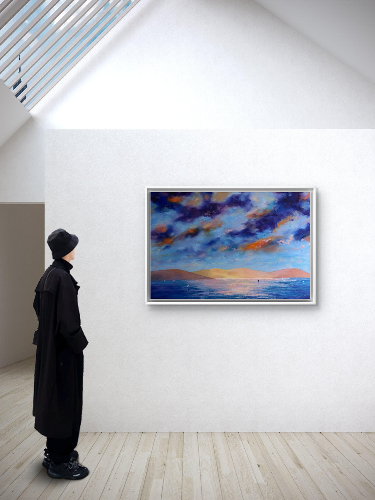 A framed landscape painting of a dramatic blue and orange sunset sky over water, displayed on a white gallery wall. A person in a long black coat and black hat stands to the left, looking at the artwork in a bright, modern room with light wood floors.