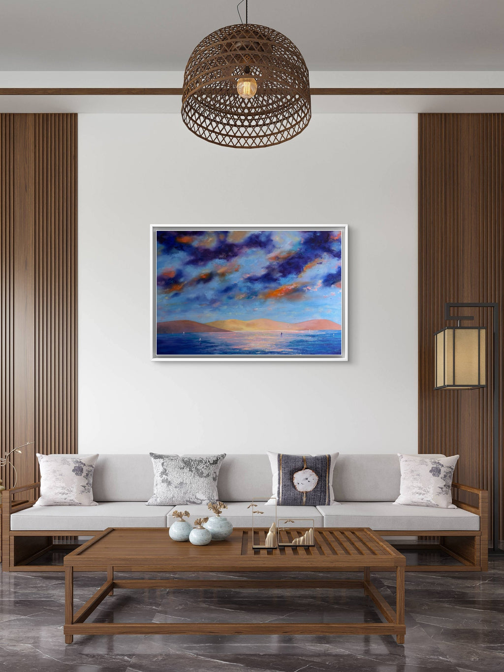 A framed landscape painting of a dramatic blue and orange sunset sky over water, displayed on a white wall in a modern living room setting with a wooden sofa, coffee table, and pendant light.